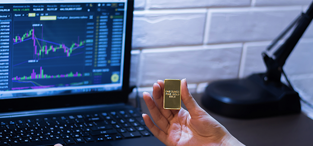 a female hand holding a little bar of gold in front of a laptop with technical charts referring to trading gold forex