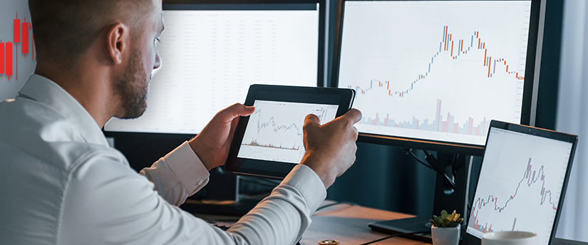 A trader holding a tablet with multiple data-filled monitors in the background, highlighting the role of trading psychology.