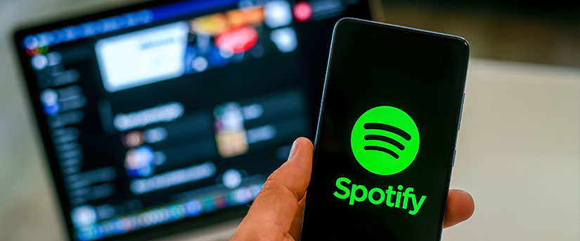 A user holds a mobile phone with the Spotify logo, with a laptop in the background, capturing a multimedia experience.