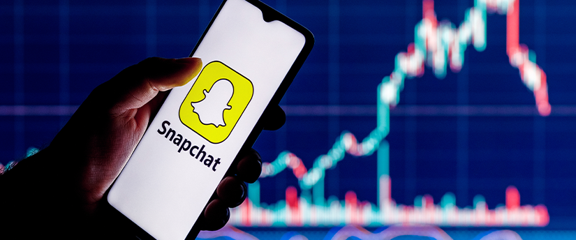 A user holding a mobile phone displaying the Snapchat app icon, with financial charts in the background.