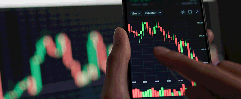 A mobile trader holding a cell phone with a CDS stock chart on it in front of a computer screen.