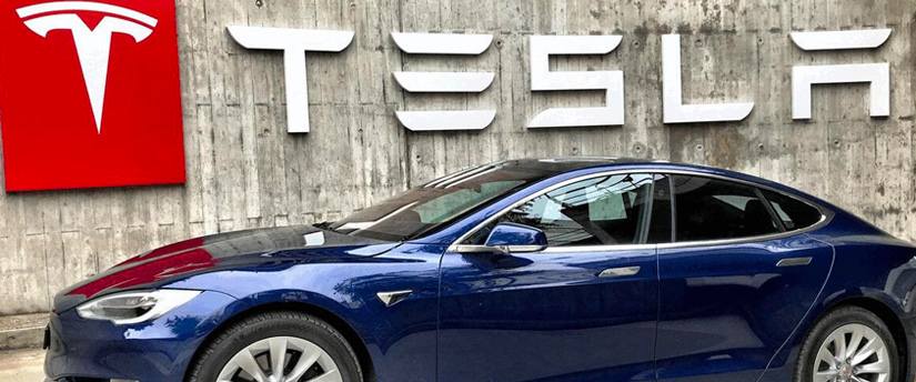A Tesla car parked under the iconic Tesla sign, symbolizing the electric vehicle manufacturer.