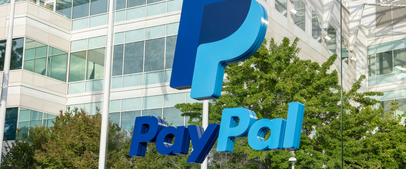 A modern glass building with the PayPal logo on top, surrounded by trees and parked cars.