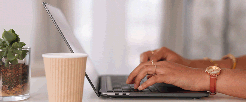 A woman focused on her laptop, typing intently while enjoying a cup of coffee beside her.