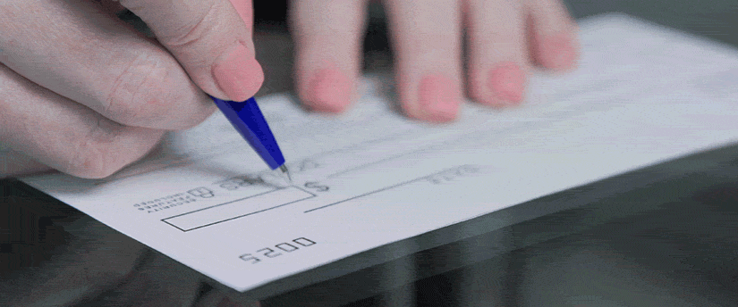 A woman's hand poised over a checkbook, carefully writing a check, with a pen in hand and a soft background.

