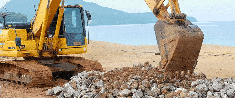 A yellow excavator is actively digging up rocks on a sandy beach, showcasing its powerful machinery in action.