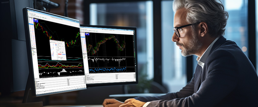 A suited man works at a desk with two monitors, each showing different trading platforms and market information.