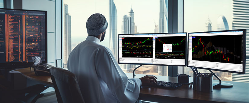 A man wearing a turban sits at a desk, focused on three computer monitors displaying various tasks.