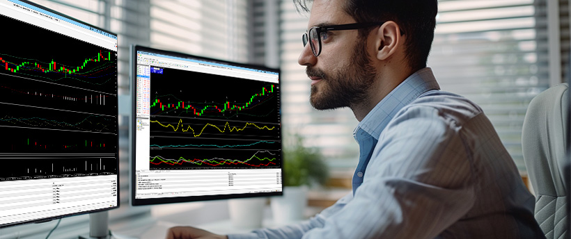A man wearing glasses analyzes forex trading data displayed on two computer screens.